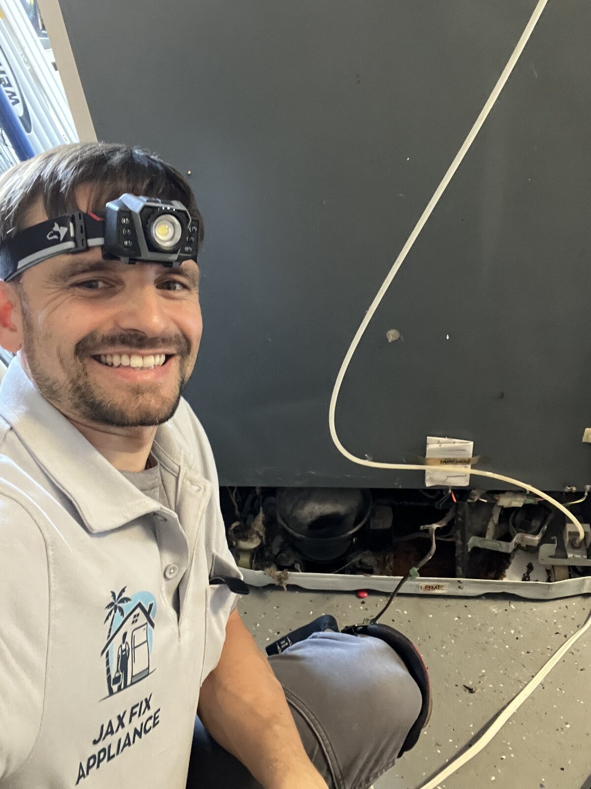 Konstantin in branded Jax Fix Appliance polo working on a refrigerator with exposed wiring